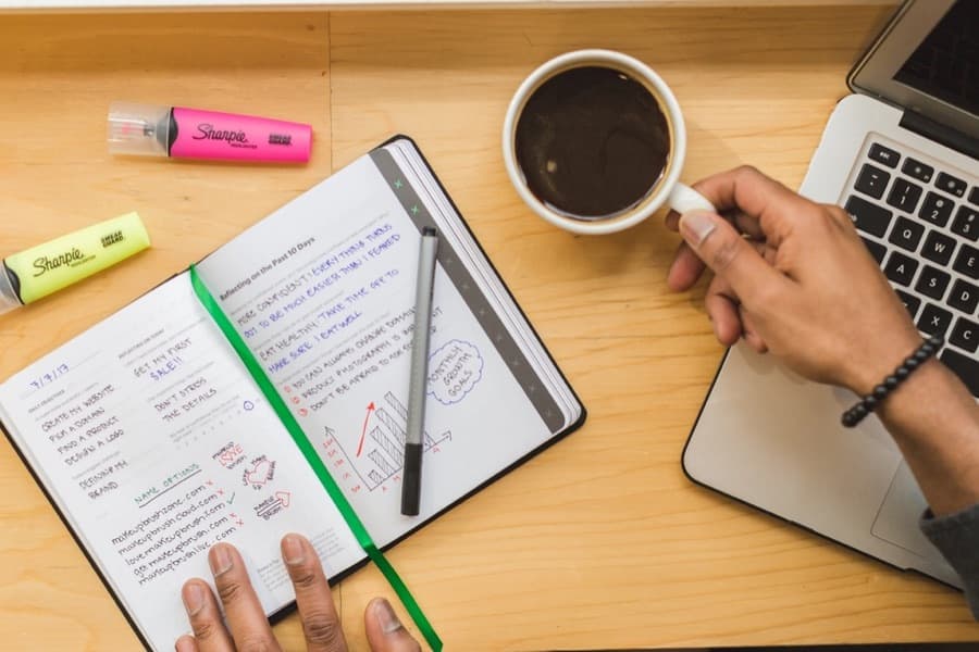 Hands writing in a notebook next to a cup of coffee and a laptop