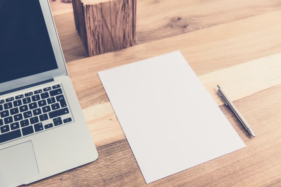 A laptop next to a blank sheet of paper and a pen on a desk
